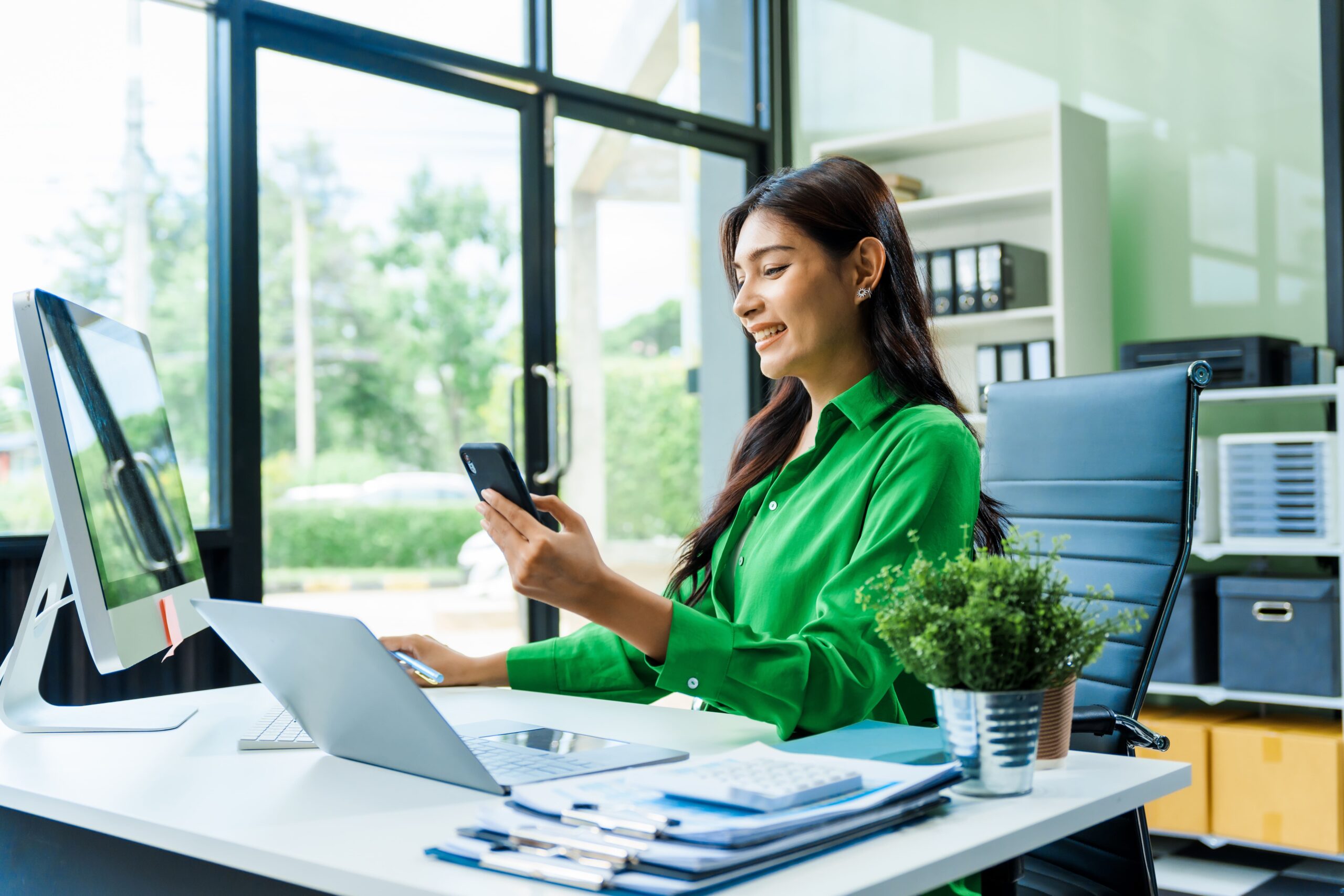 A woman using construction project management software on her smartphone while sitting in front of her desktop computer in a green shirt